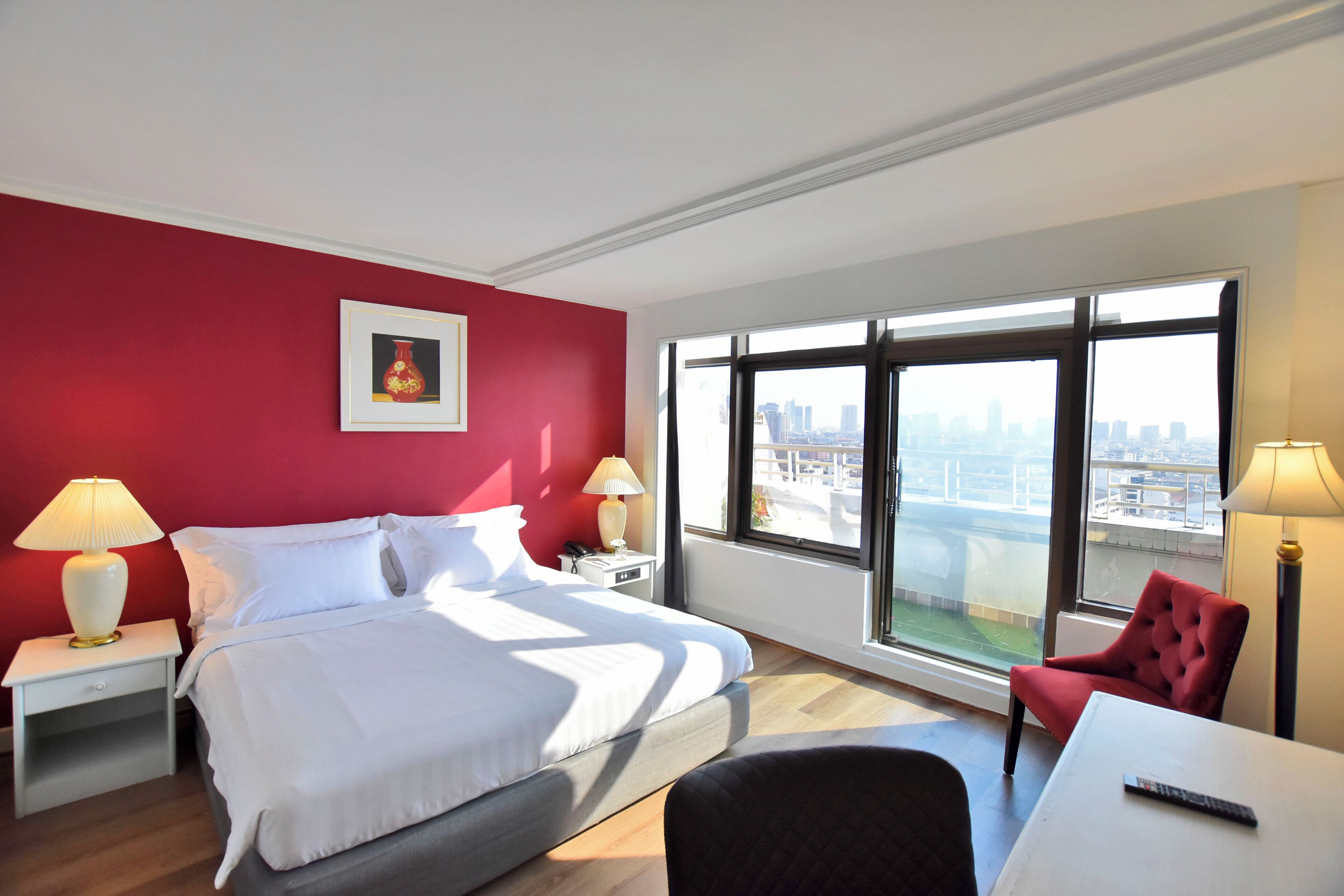 A hotel room with a bed, two bedside tables with lamps, a red chair, a small table with a remote, and a large window with a city view.
