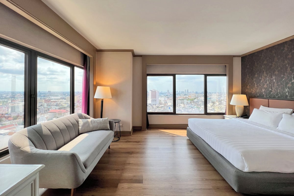 Une chambre d’hôtel avec un lit, un canapé, deux lampes, une vue sur la ville et une grande fenêtre.