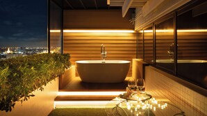 Un baño moderno con una bañera independiente, una mesa de vidrio con una copa de vino y vistas a la ciudad de noche.