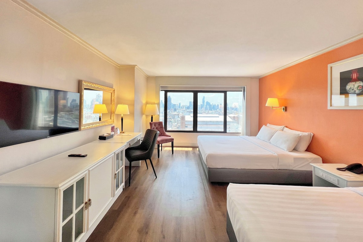 Une chambre d’hôtel équipée d’un bureau, de deux lits, d’une grande fenêtre donnant sur la ville et d’un téléviseur fixé au mur.