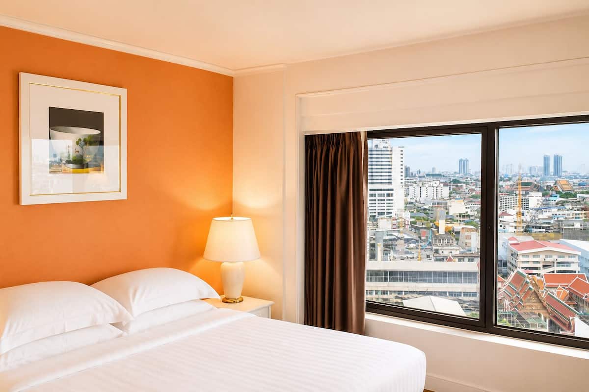 Une chambre d’hôtel avec un lit, une table de chevet, une lampe, un tableau encadré et une grande fenêtre donnant sur la ville.