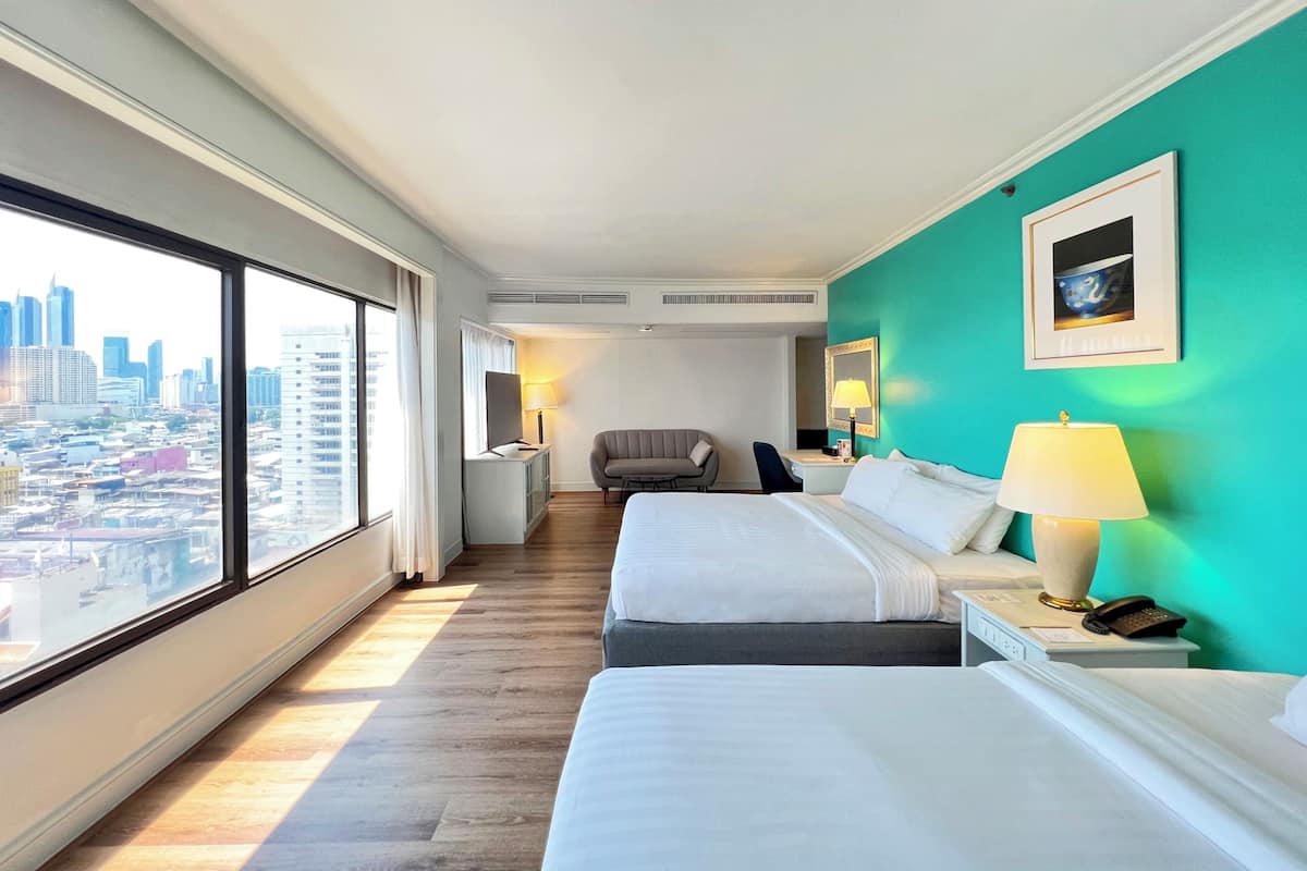 Une chambre d’hôtel avec deux lits, un bureau, une chaise, une grande fenêtre donnant sur la ville et un mur d’accent de couleur turquoise.