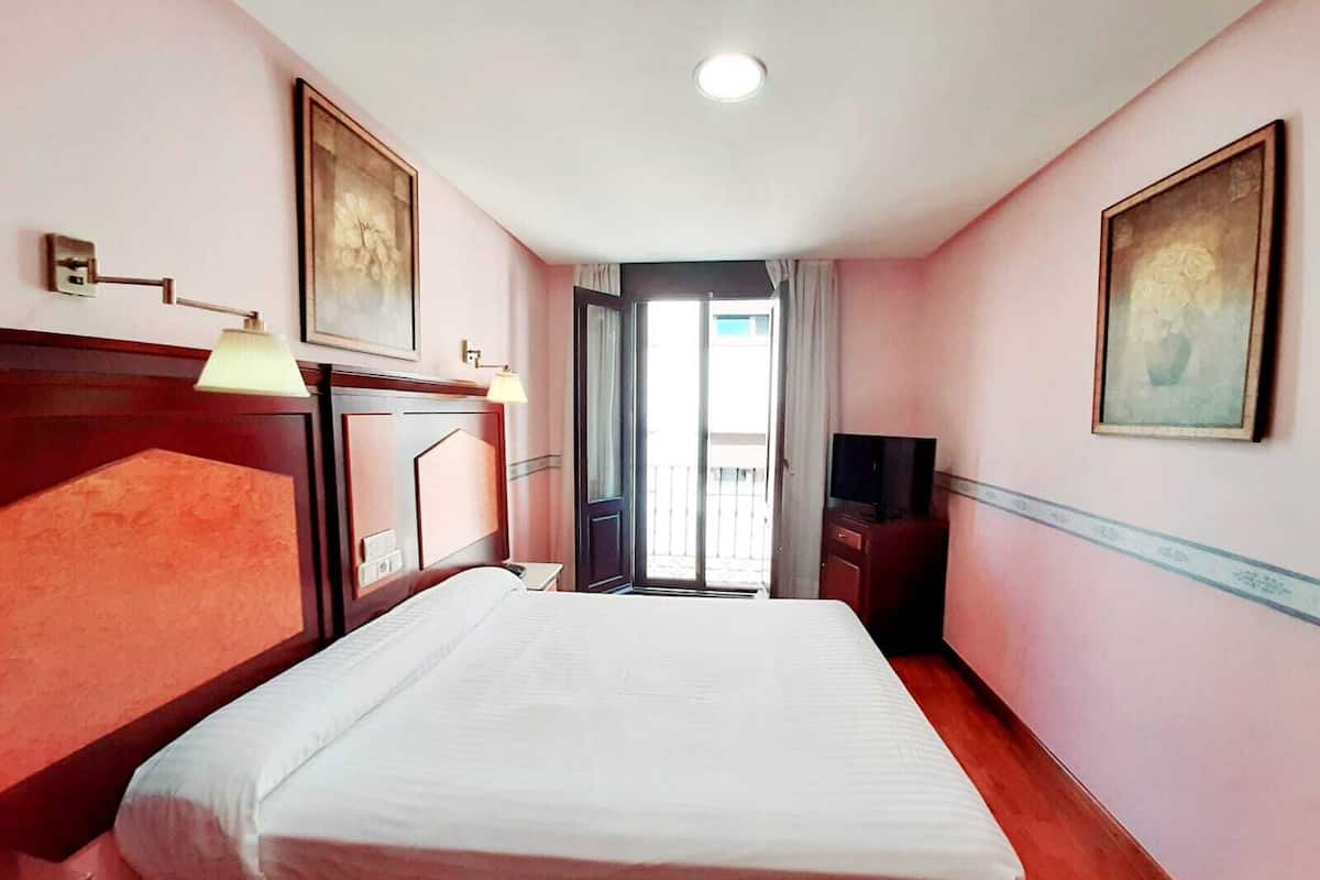 A hotel room with a large bed, a desk, a television, and a balcony with curtains.