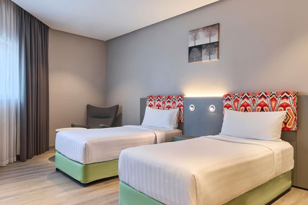 Habitación de hotel con dos camas, un escritorio y una silla. Se observa una ventana con cortinas, una obra de arte en la pared y un cabecero con diseño estampado.