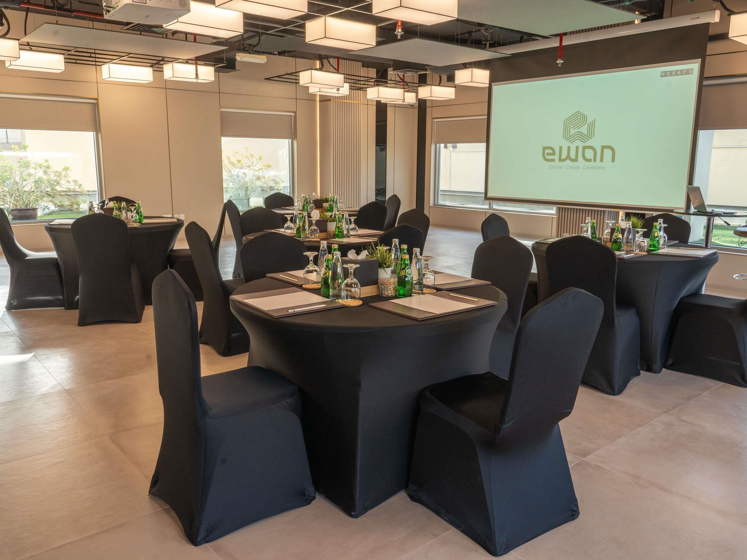 Un salón de conferencias con mesas redondas, sillas negras y una pantalla de proyector que muestra el logo 'ewan'.