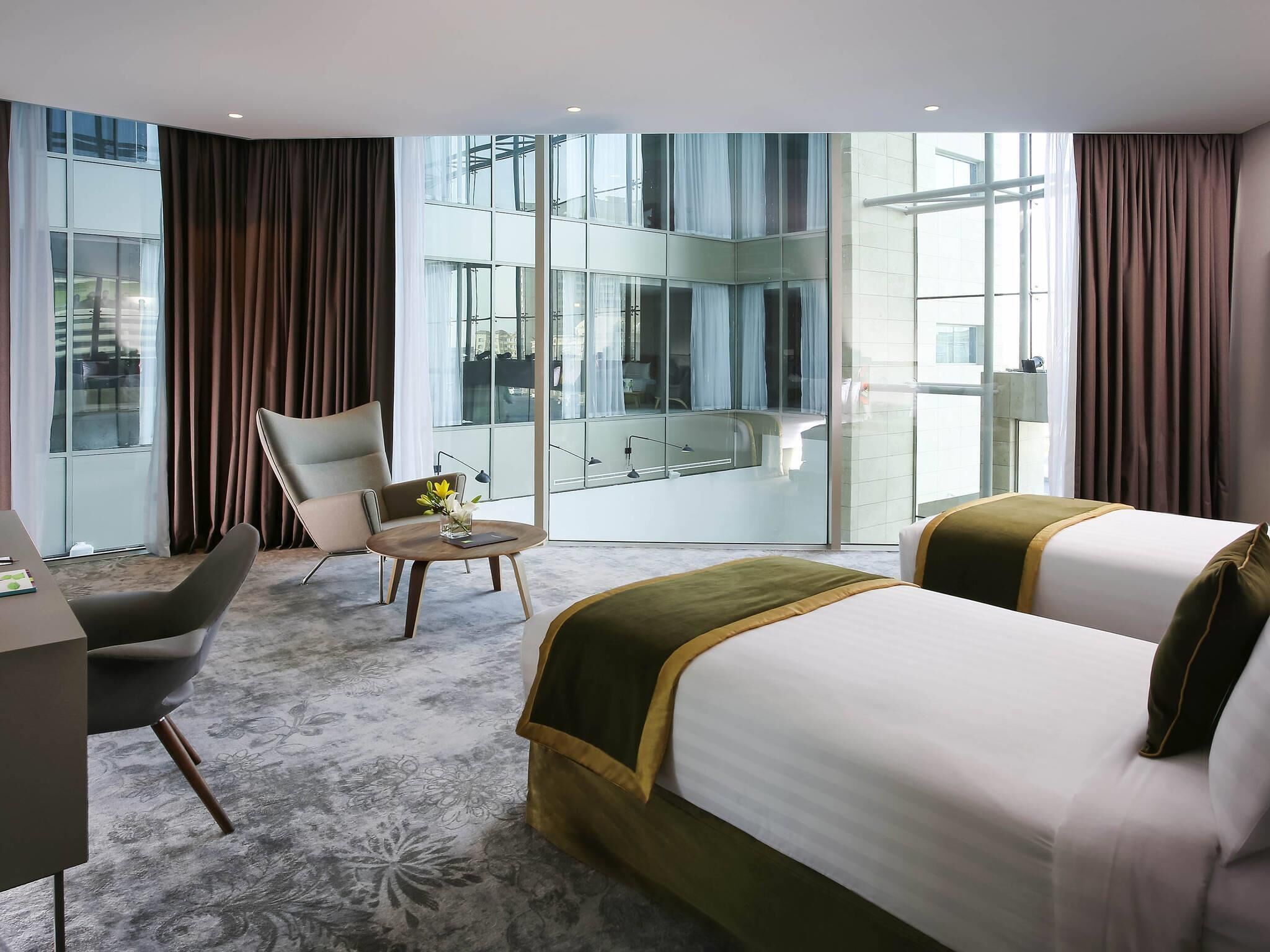 A hotel room with a bed, a desk, a chair, and a view of a building through large windows.