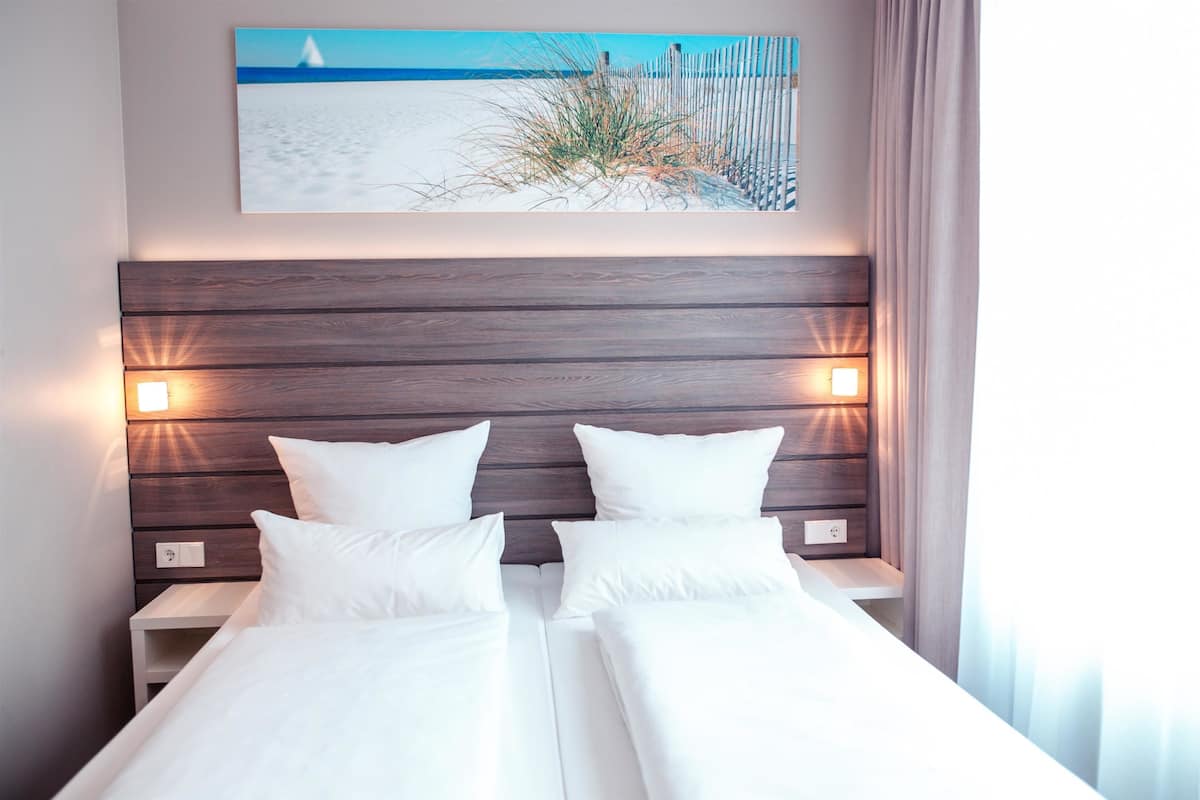 Cama de casal com lençóis brancos, cabeceira de madeira, abajures laterais e um quadro com tema praiano na parede.