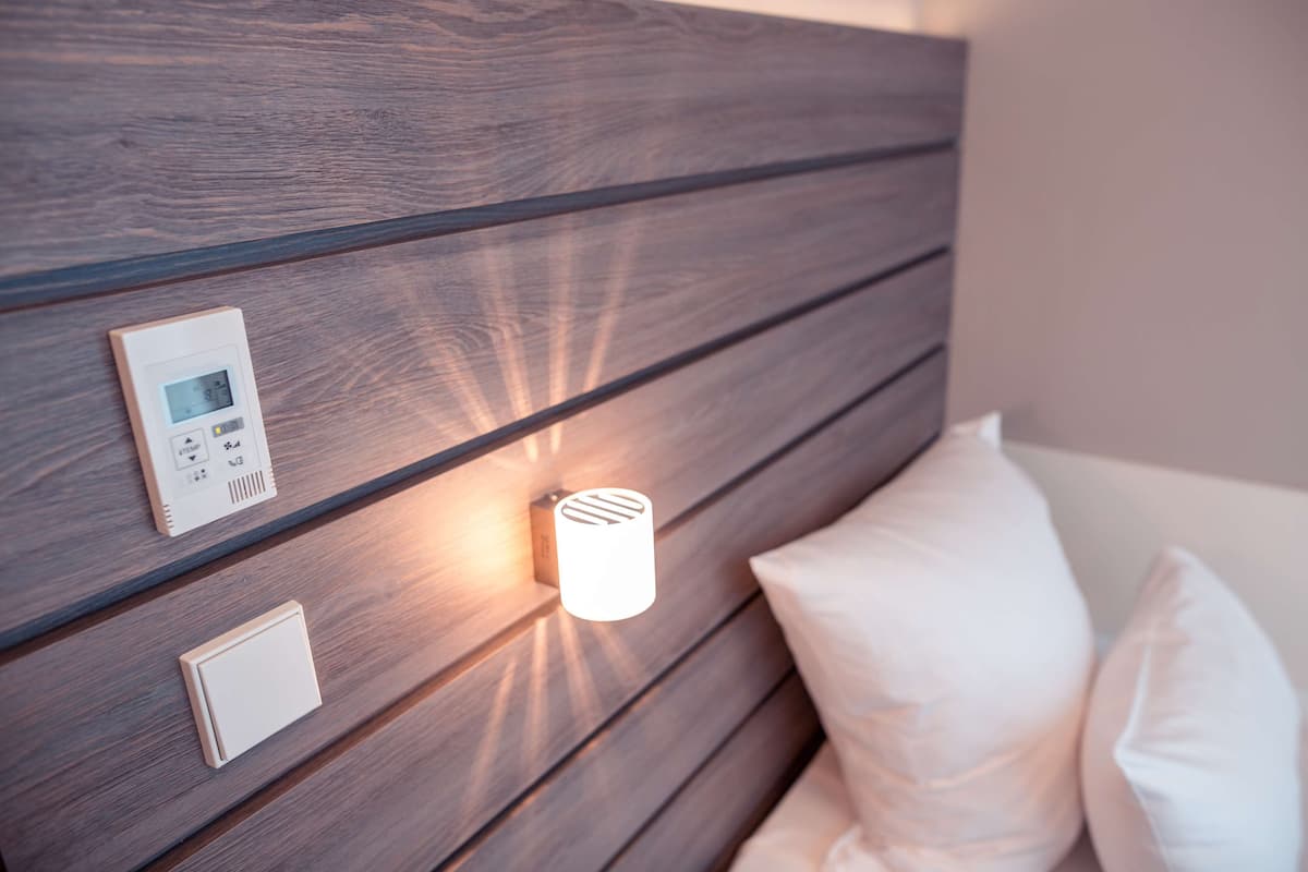 Quarto moderno com cabeceira de madeira, painel de controle na mesa de cabeceira, luminária fixada na parede e travesseiro branco.