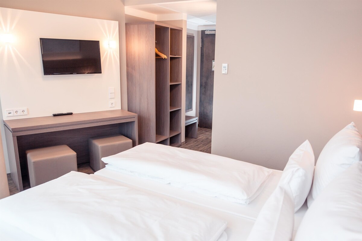 Quarto de hotel com cama, escrivaninha com televisão, armário e chuveiro.