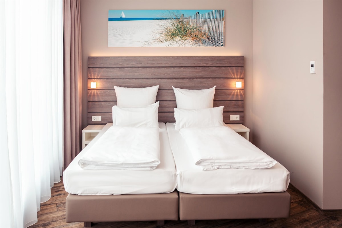 Uma cama de casal com roupa de cama branca, cabeceira de madeira, abajures e uma pintura com tema praiano acima.