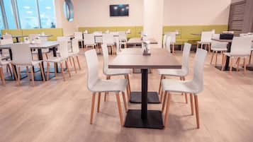 Uma sala de jantar moderna com várias mesas e cadeiras, uma TV de tela plana na parede e grandes janelas que oferecem luz natural.