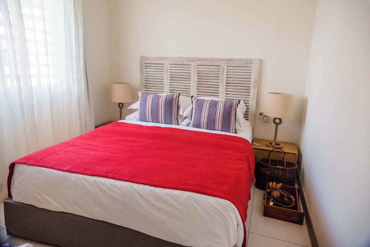 Un lit avec une couverture rouge, deux oreillers, une tête de lit en bois, deux lampes de chevet et une petite commode en bois avec des paniers.