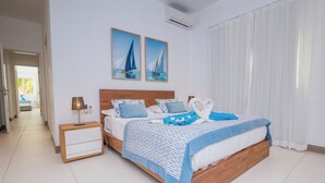 Een slaapkamer met een houten bed, wit- en blauwe beddengoed, twee schilderijen van zeilboten, een nachtkastje met een lamp, en een raam met witte gordijnen.