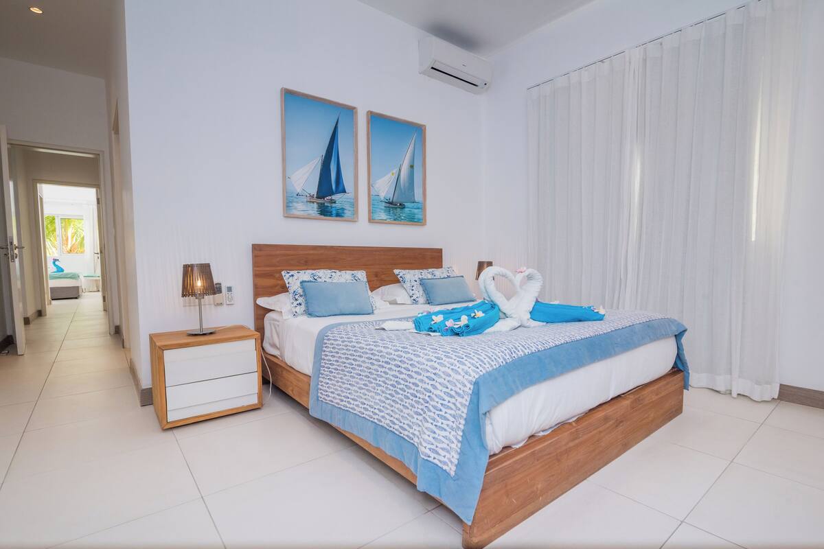 Une chambre avec un lit en bois, une literie blanche et bleue, deux tableaux représentant des voiliers, une table de chevet avec une lampe et une fenêtre avec des rideaux blancs.