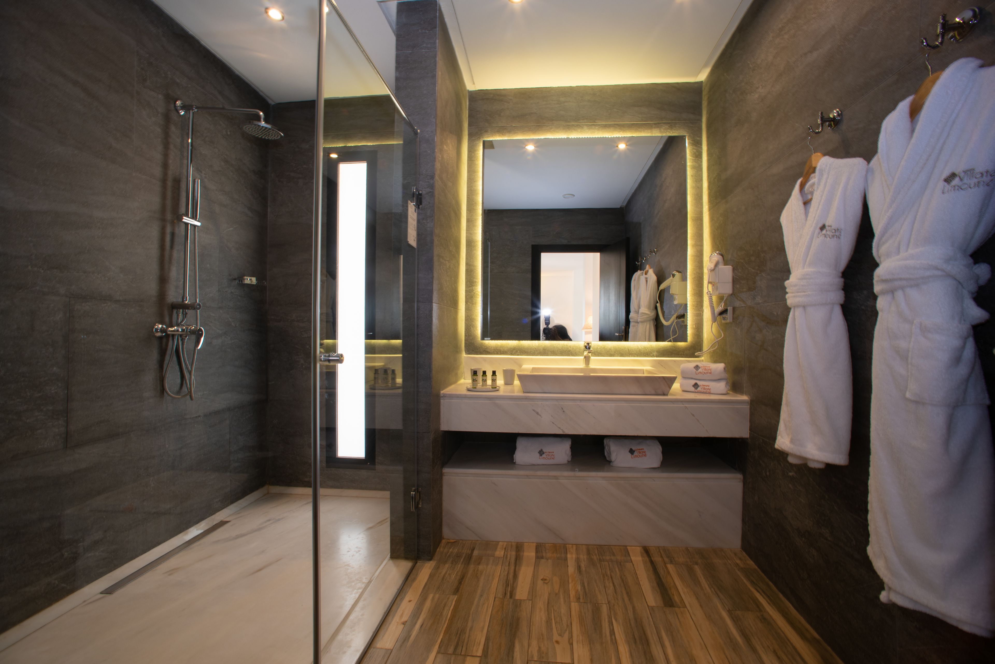 A modern bathroom with a glass-enclosed shower, a large mirror, and two white robes hanging on hooks.
