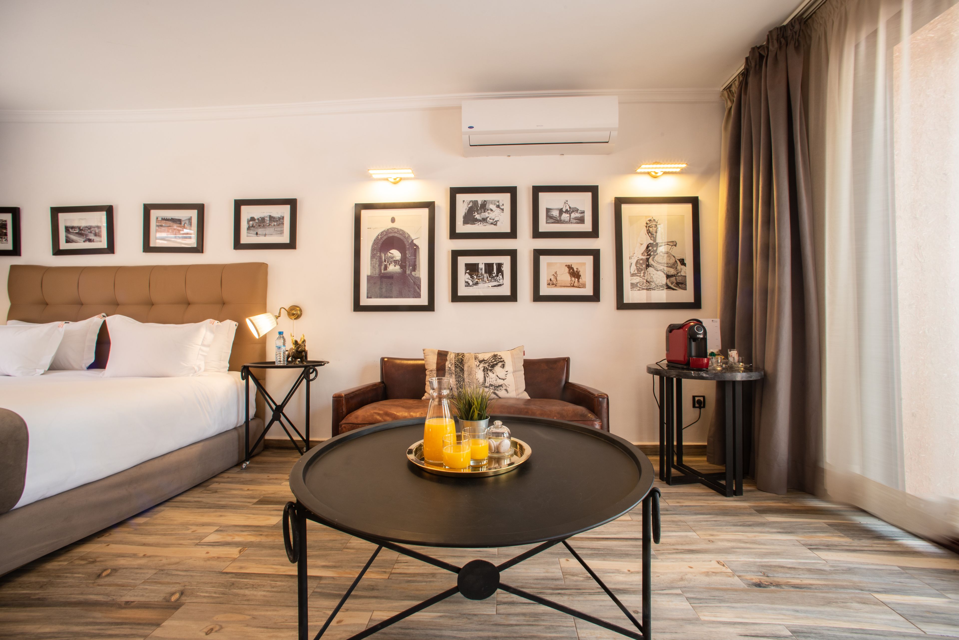 A hotel room with a bed, a round table with a tray of orange juice, a coffee machine, and framed pictures on the wall.