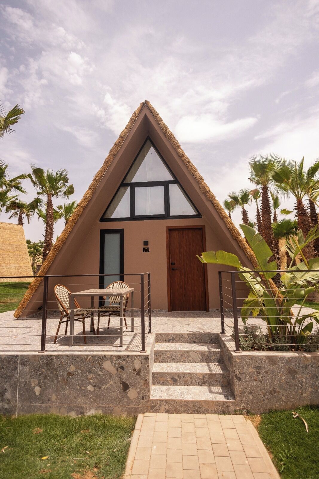 A triangular building with a thatched roof, a wooden door, and a small outdoor seating area with chairs and a table.