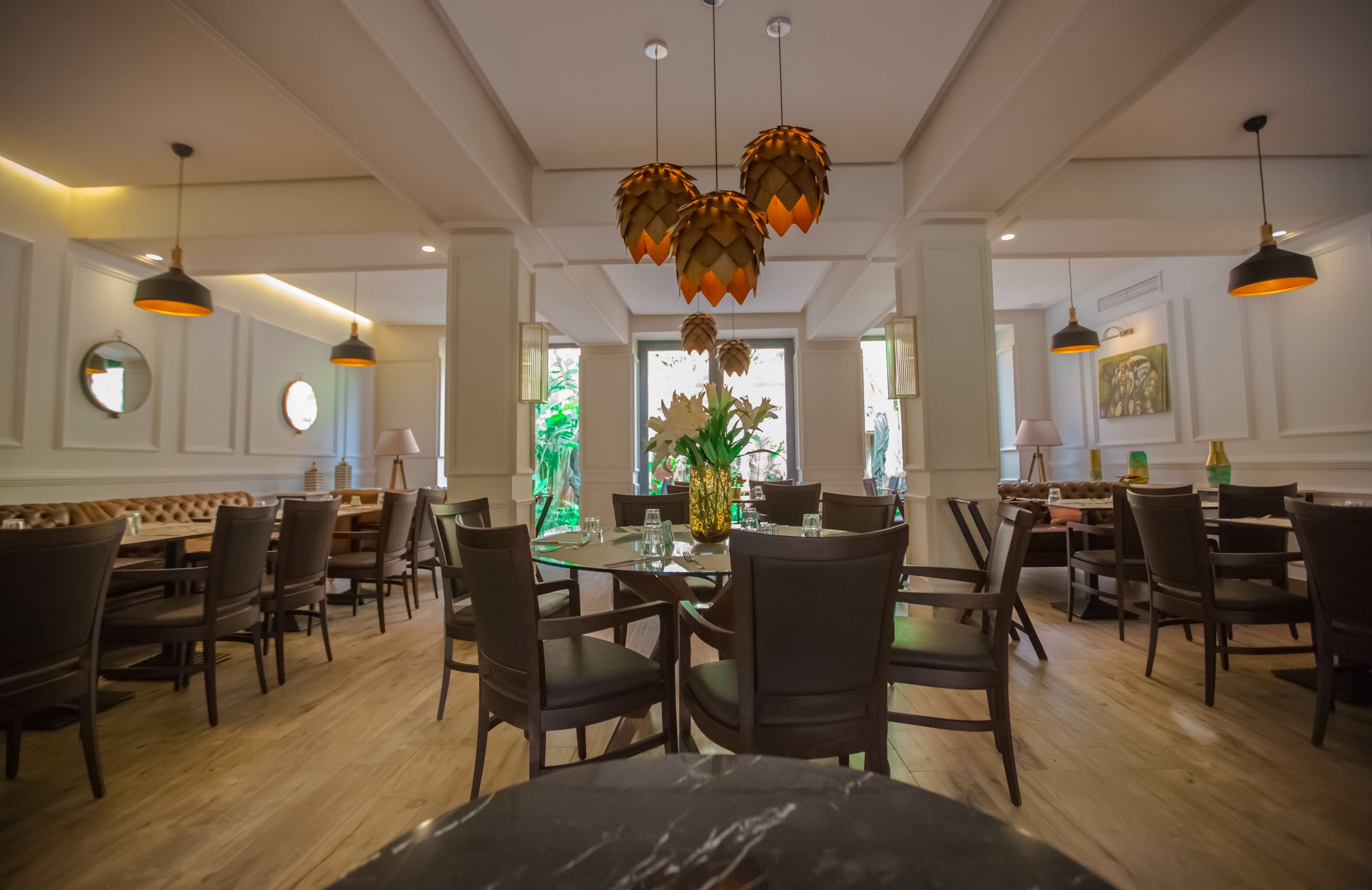 A dining area with wooden tables and chairs, hanging pendant lights, and a central floral arrangement.