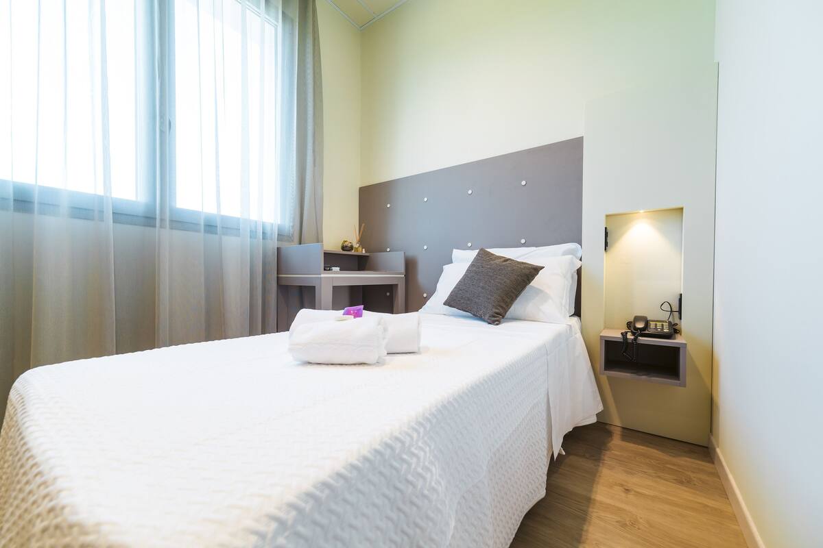 Una cama bien hecha con ropa de cama blanca, una mesita de noche con una lámpara y una pequeña estantería con un teléfono.