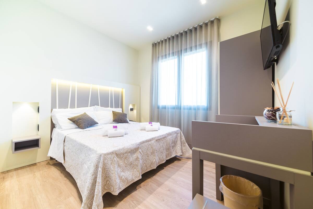Una habitación de hotel con una cama, un televisor en la pared y un ventanal con cortinas.