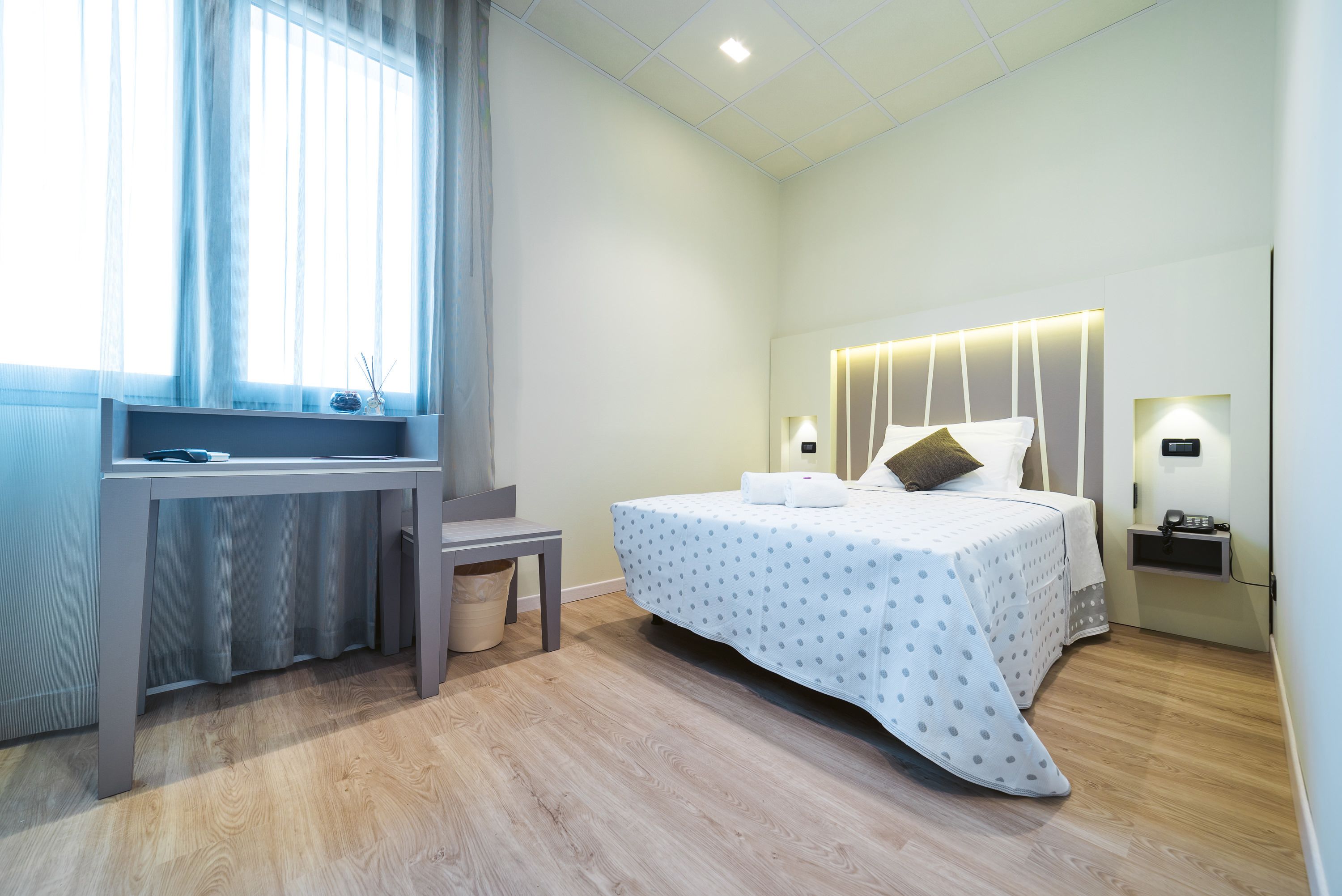 A modern hotel room with a bed, bedside table, lamp, and a desk with a chair.
