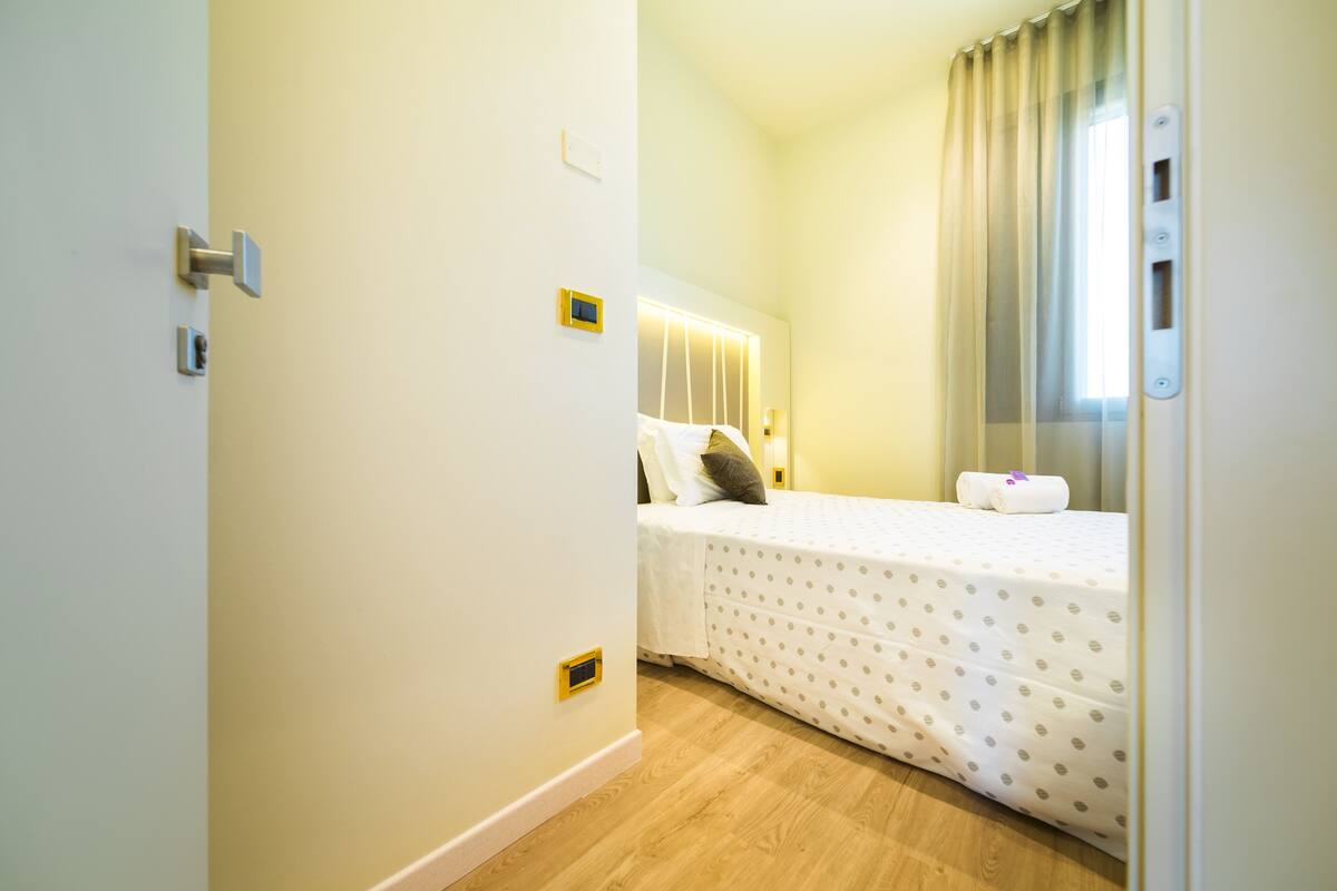 Una habitación de hotel con cama, mesita de noche, ventana con cortinas y puerta con pomo.