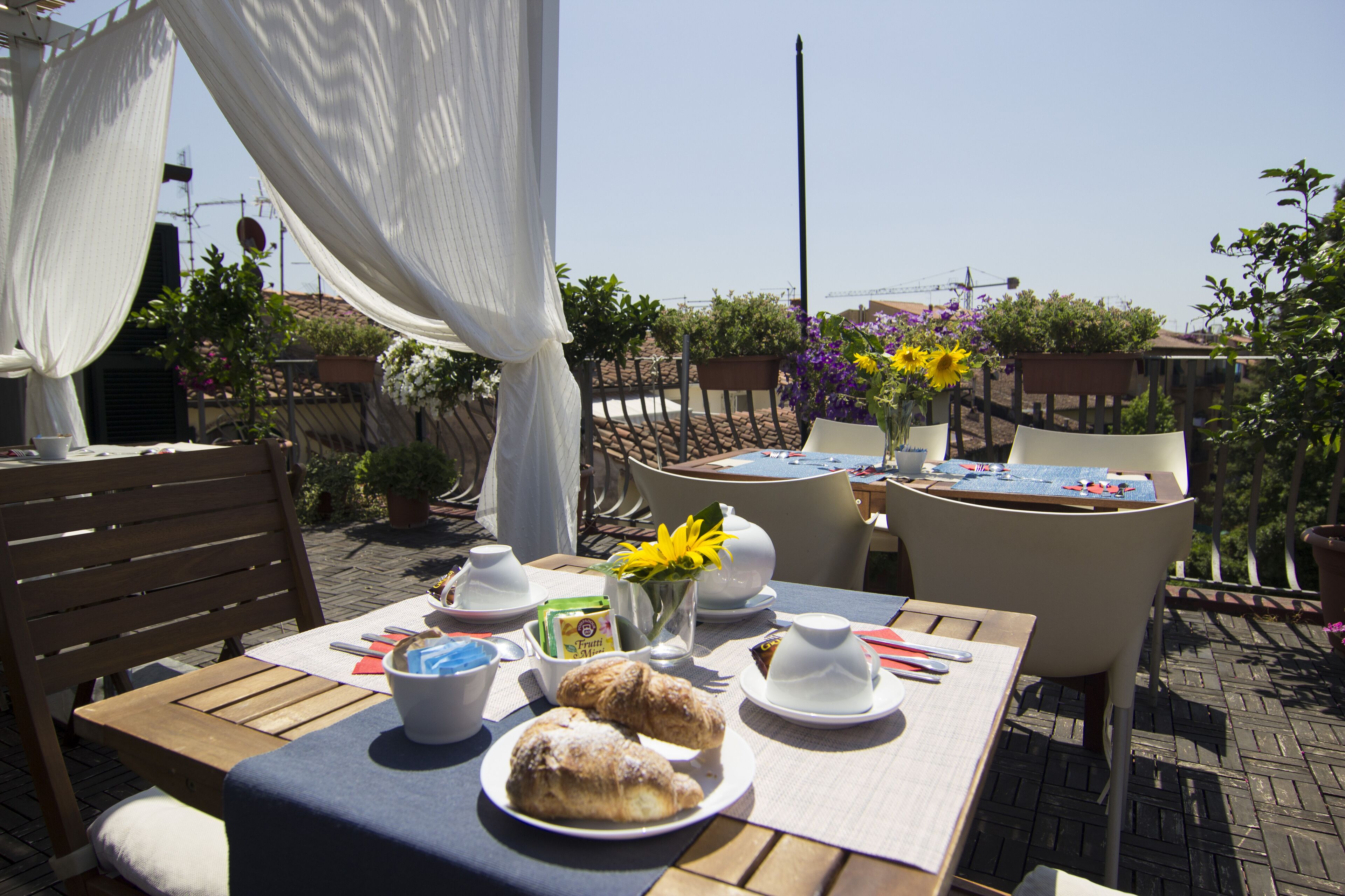 Un área de comedor al aire libre con una mesa preparada para dos personas, que incluye pasteles, un florero con flores y vistas del jardín circundante y edificios.