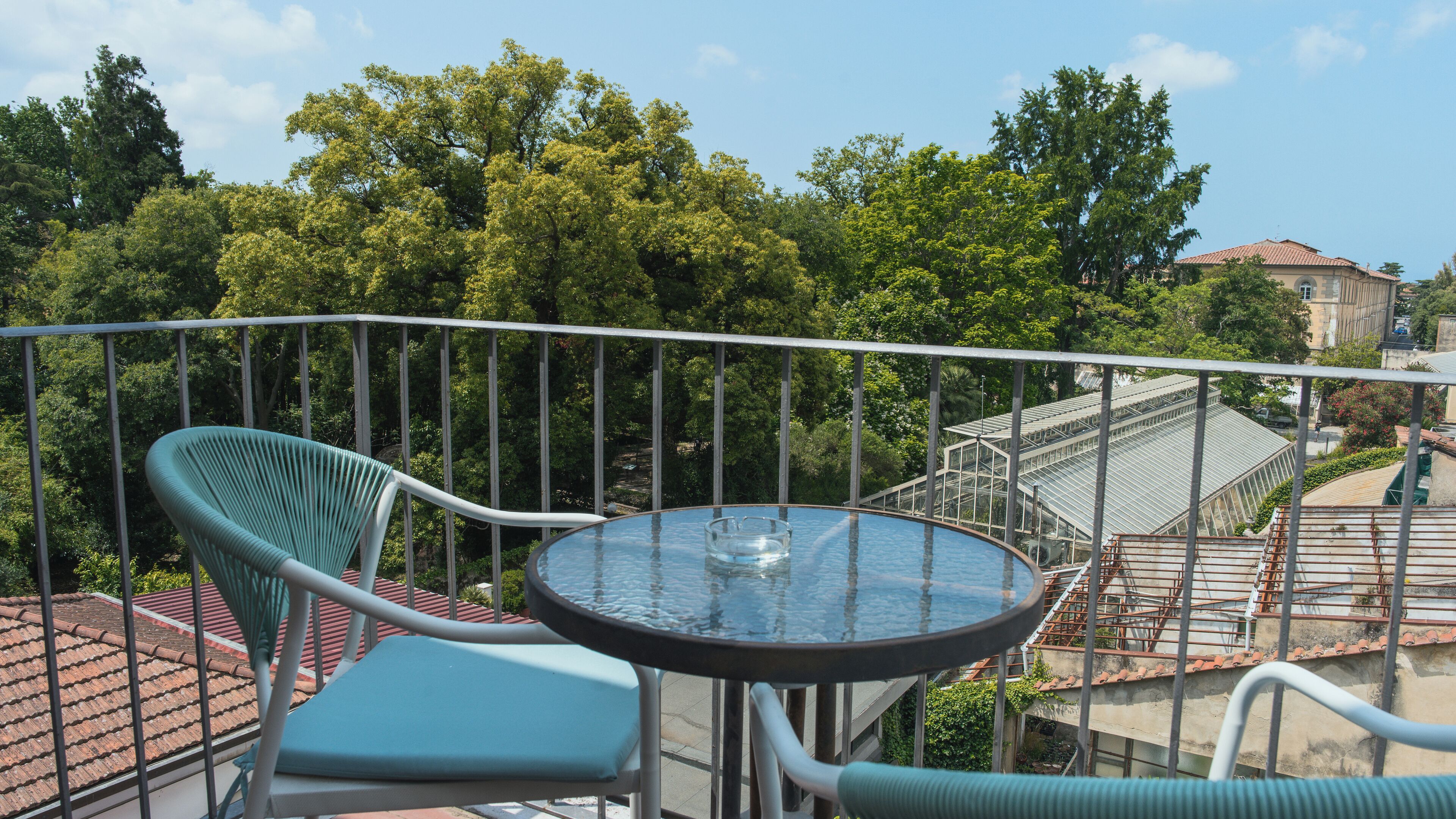 Ein Balkon mit Glastisch und zwei Stühlen, mit Blick auf einen Garten und Gebäude.