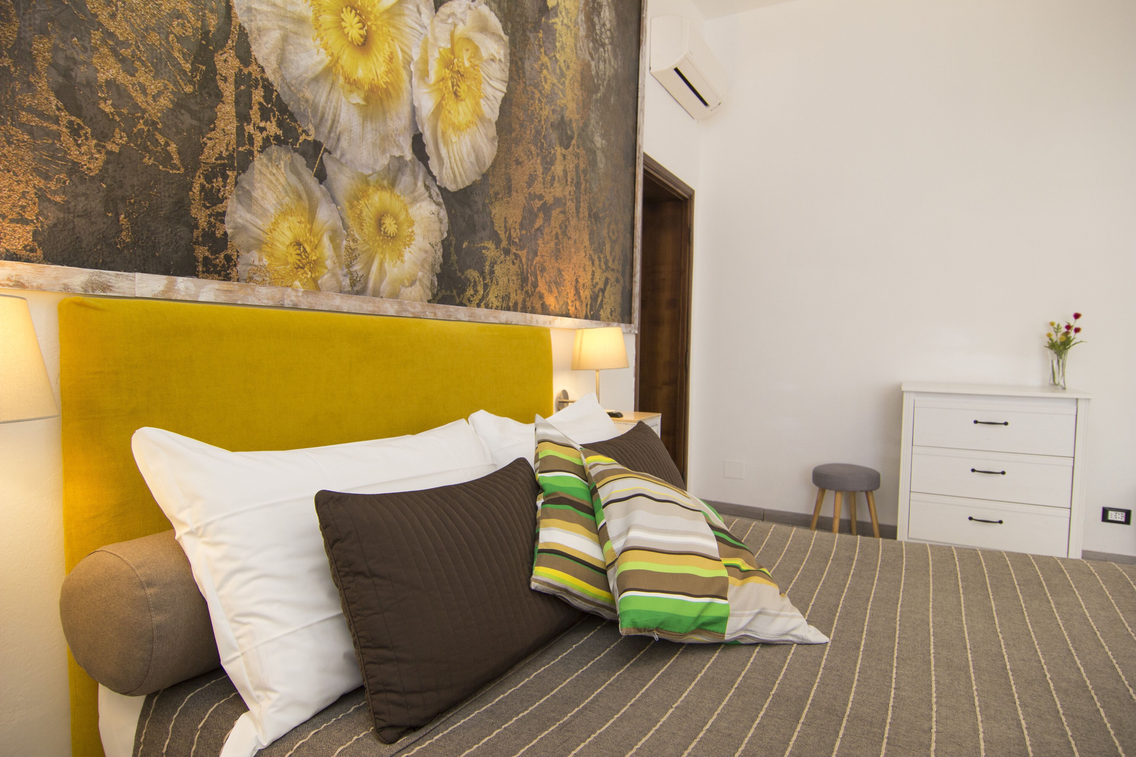 Una cama bien hecha con ropa de cama a rayas y varios almohadones, una mesita de noche blanca con cajones, y una pared decorada con un cuadro floral.