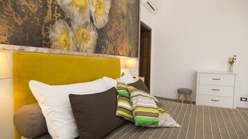 Una cama bien hecha con ropa de cama a rayas y varios almohadones, una mesita de noche blanca con cajones, y una pared decorada con un cuadro floral.