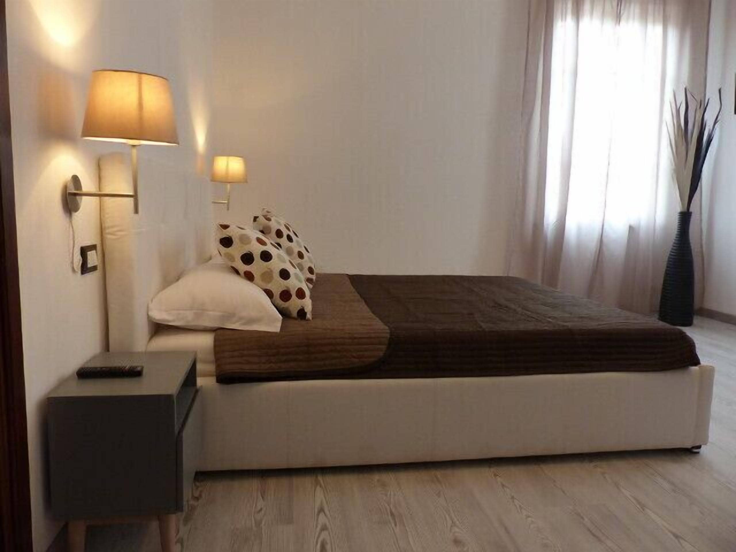 Un dormitorio moderno con una cama grande, dos lámparas de noche, una mesita de noche y un florero con plantas altas.