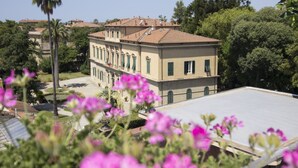 A two-story building with a tiled roof, surrounded by lush greenery and palm trees. - 7 Rooms (Pisa)