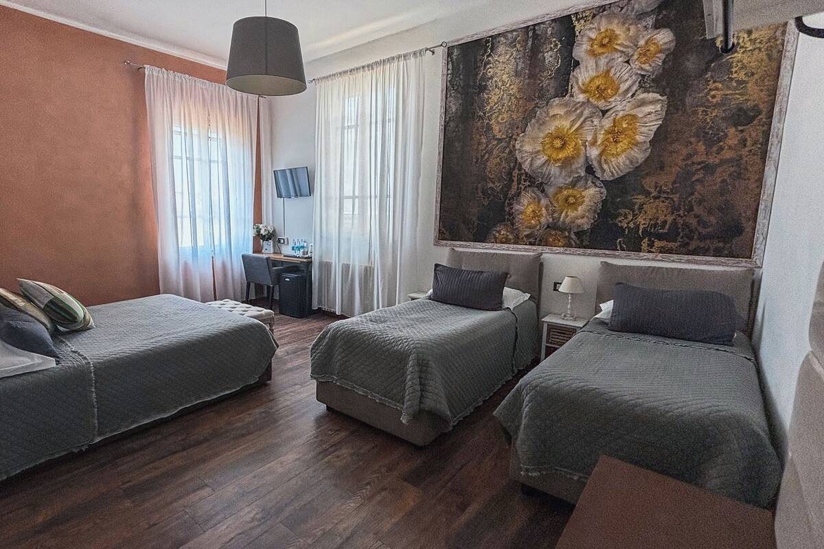 Hotellihuone, jossa on kaksi erillistä sänkyä, työpöytä tuolilla, suuri ikkuna verhoilla ja seinällä koristeellinen kukka-aiheinen taulu.