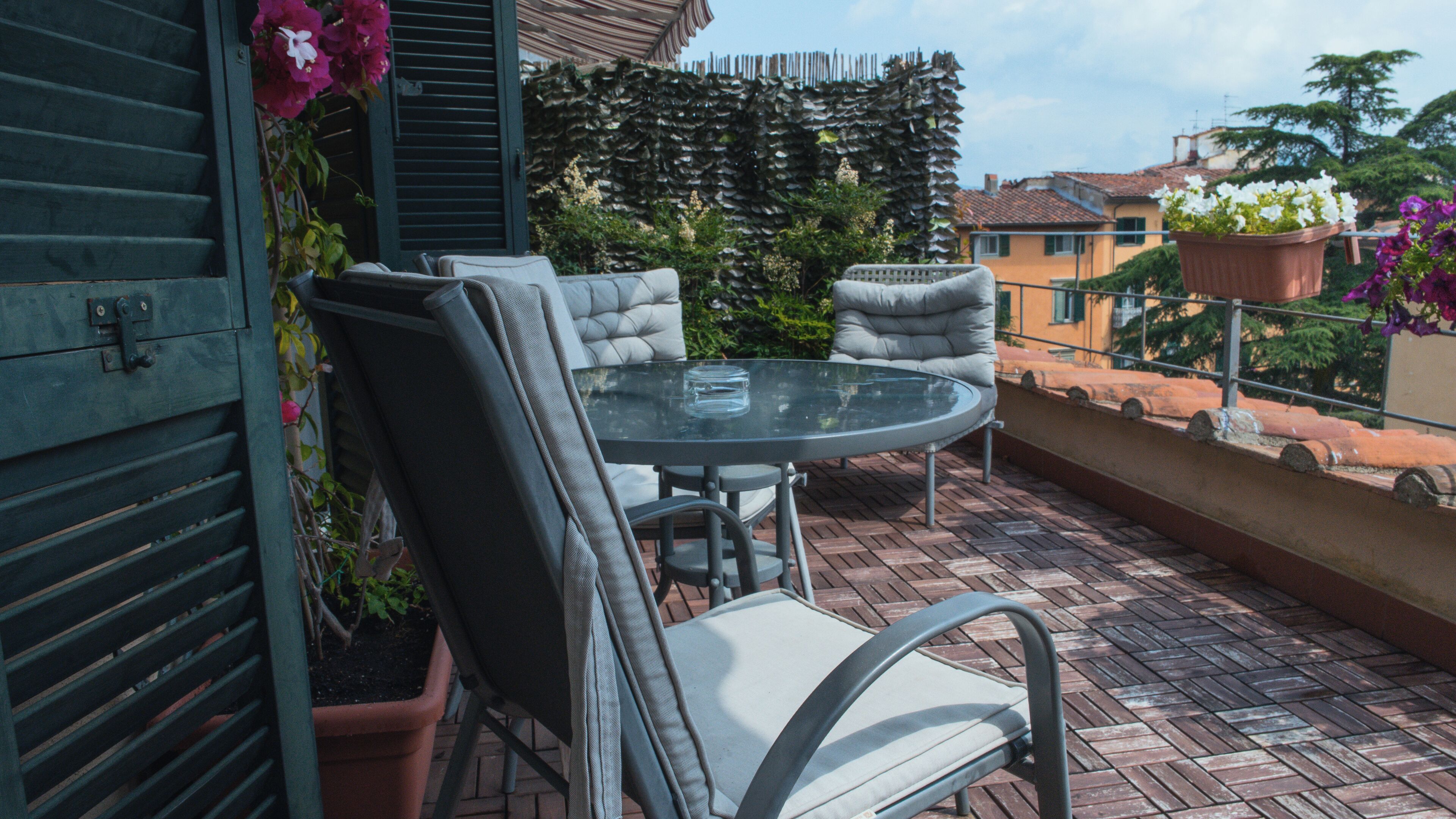 Een terras met een ronde glazen tafel en beklede stoelen, omringd door potplanten en een stenen muur.