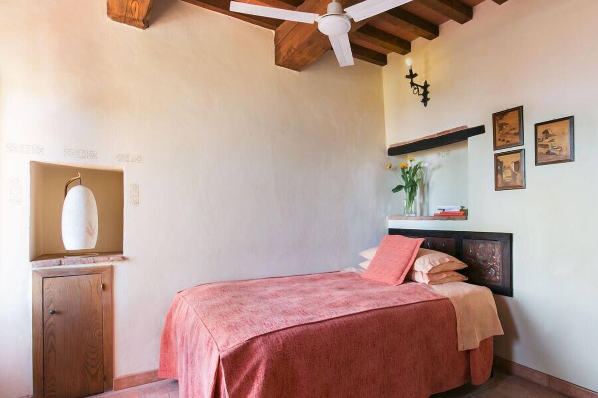 Un dormitorio con una cama, un ventilador de techo, un armario de madera y un estante en la pared con un florero y fotografías.