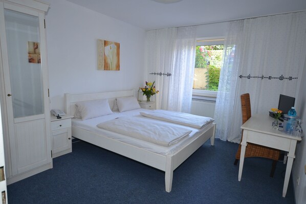 A bedroom with a double bed, a bedside table with a lamp, a chair, a desk with a computer, and a window with curtains. - Alpha Hotel Garni (Dietzenbach)