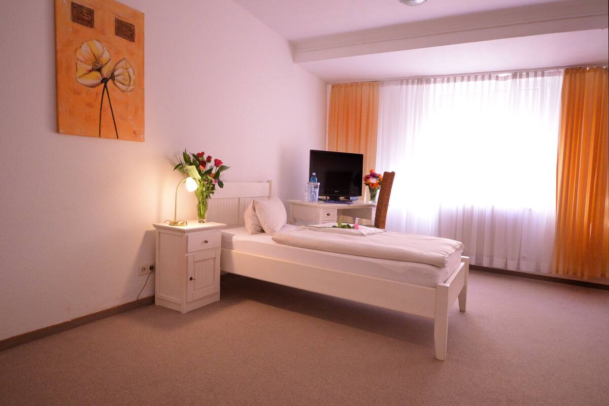 A hotel room with a single bed, a bedside table with a lamp, a TV, and a window with curtains.