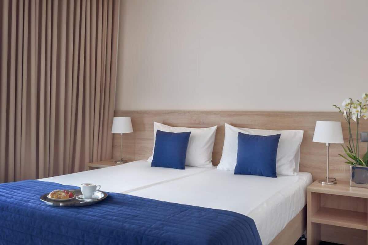 Une chambre d’hôtel avec un lit, deux tables de chevet, une lampe, un plateau avec une tasse et une pâtisserie, et un rideau.