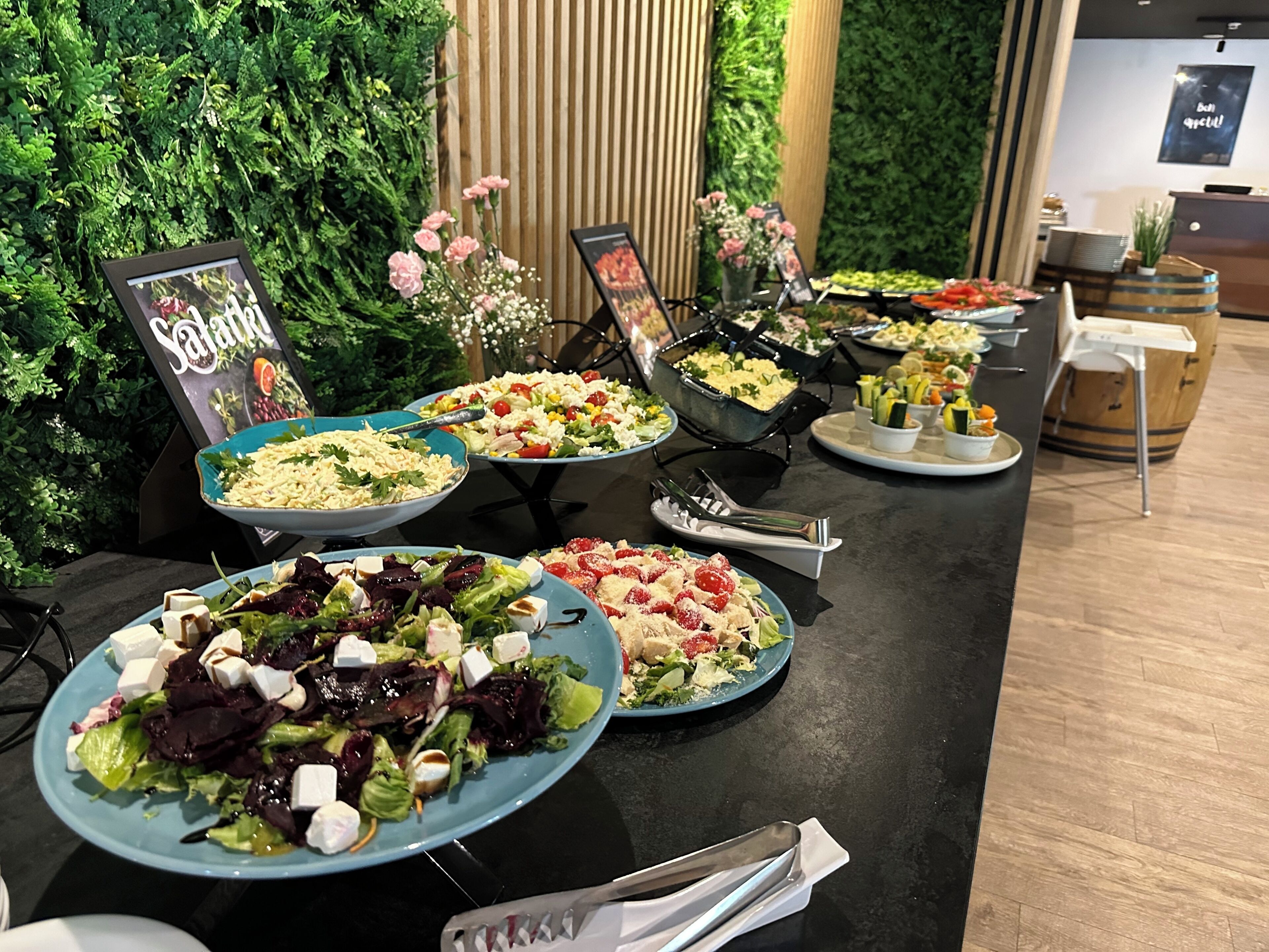 Une table de buffet garnie de différents plats, une pancarte indiquant « Saucisson », et un mur vert avec des stores verticaux en arrière-plan.