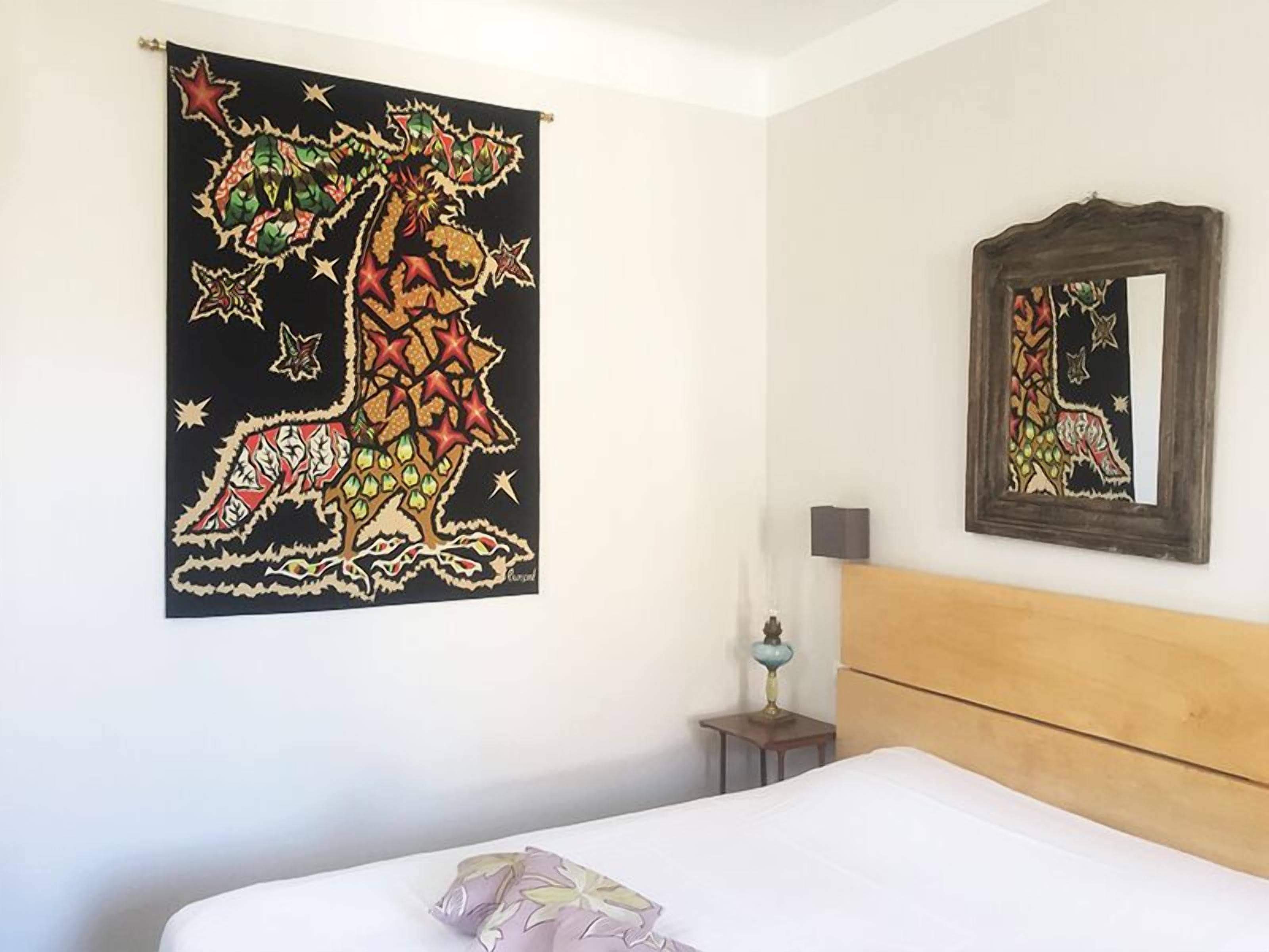 A hotel room with a bed, a nightstand, a lamp, a mirror, and a large tapestry on the wall.