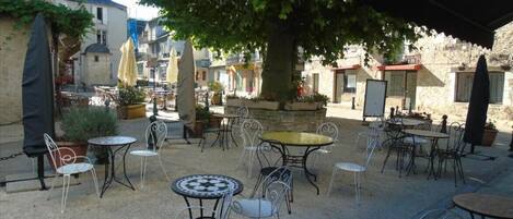 An outdoor café area with tables and chairs, surrounded by stone buildings and a large tree providing shade.