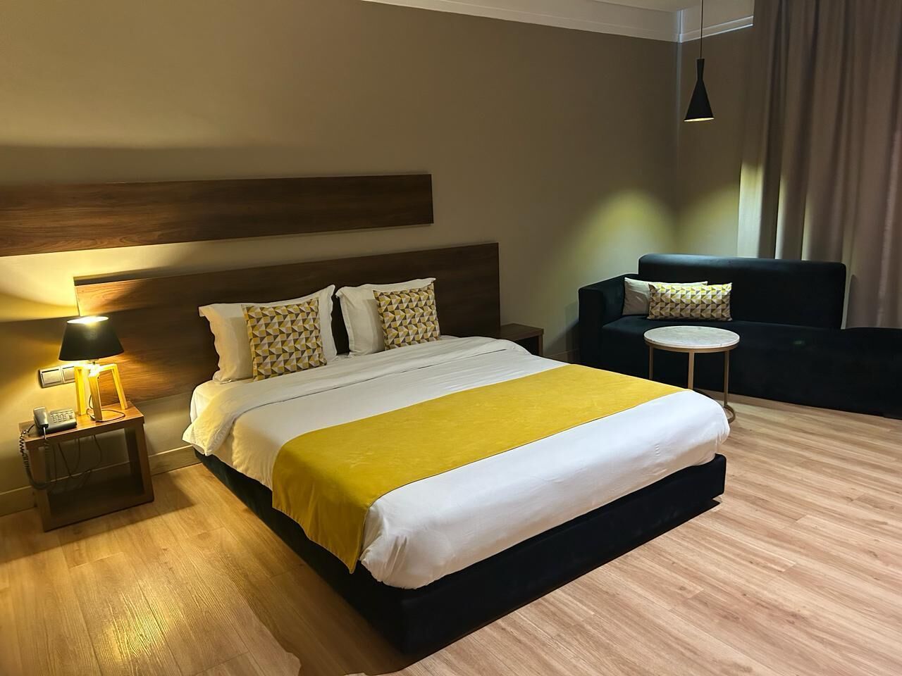 Een hotelkamer met een groot bed, nachtkastje met lamp, een klein rond tafeltje en een bank.