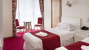 A hotel room with two beds, a small table, two chairs, a window with curtains, a door, and a bedside table with a lamp. - Afton Hotel (Eastbourne)
