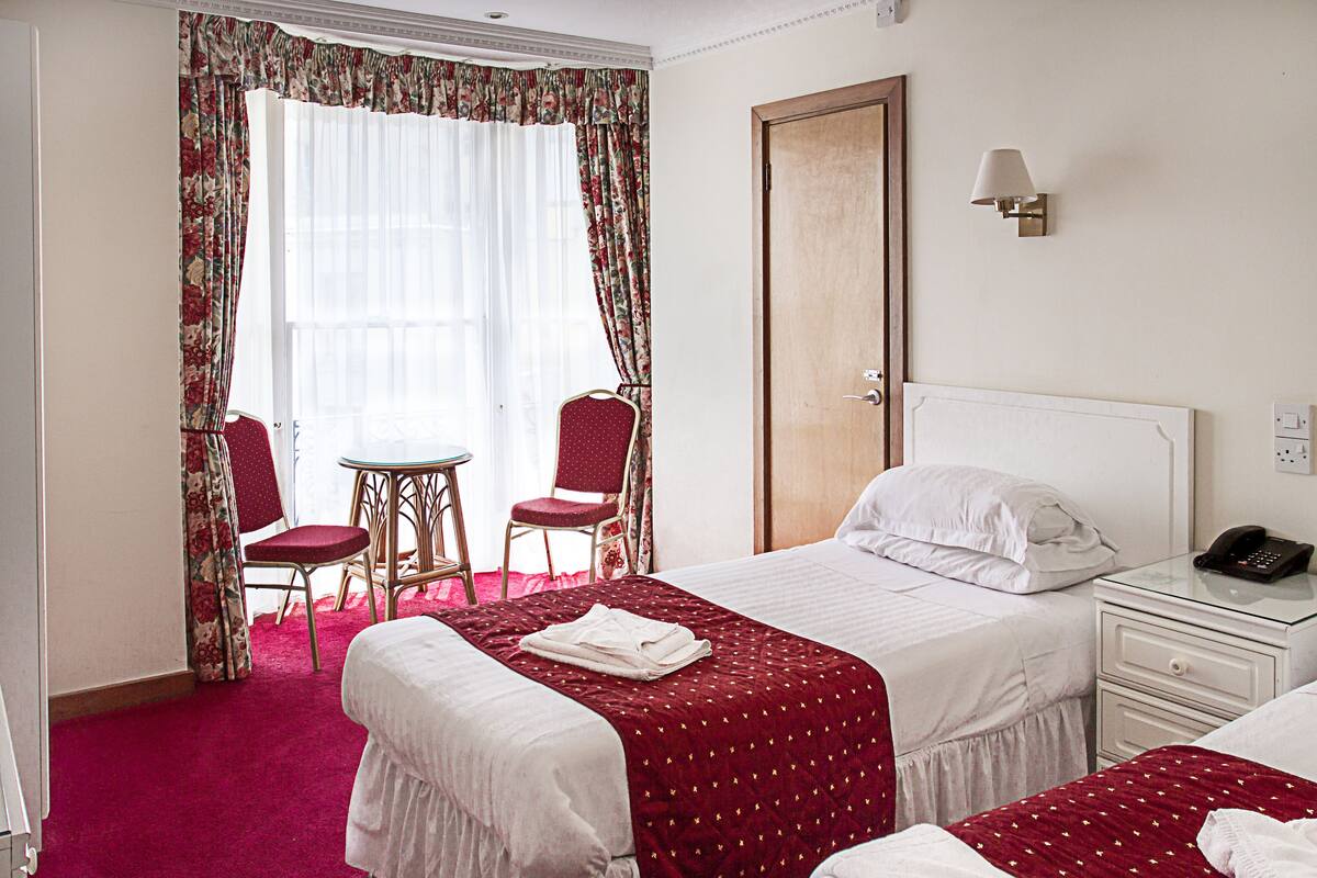 A hotel room with two beds, a small table, two chairs, a window with curtains, a door, and a bedside table with a lamp.