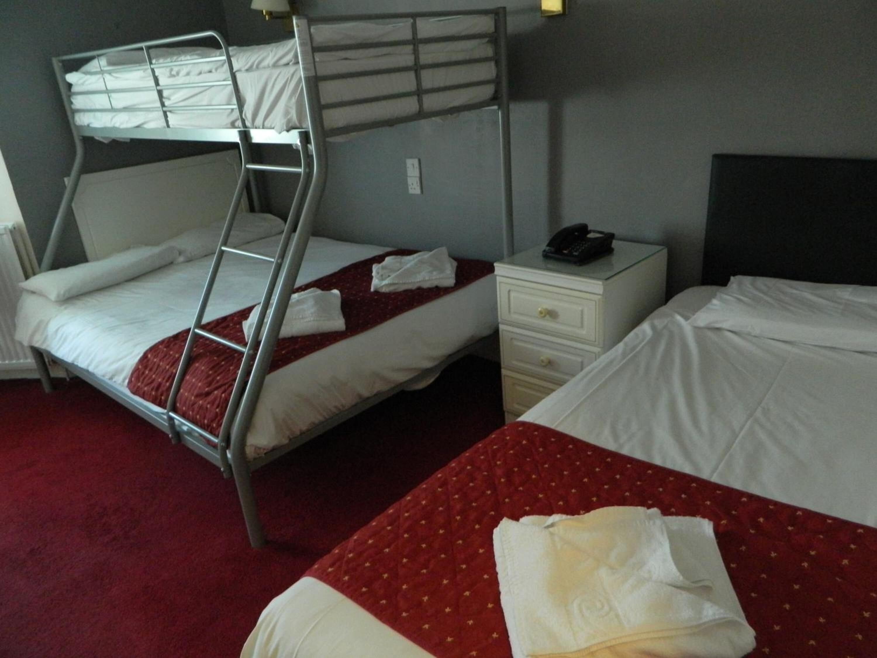 A hotel room with two beds, one with a bunk bed and the other with a single bed. There is a nightstand with a phone and a lamp.