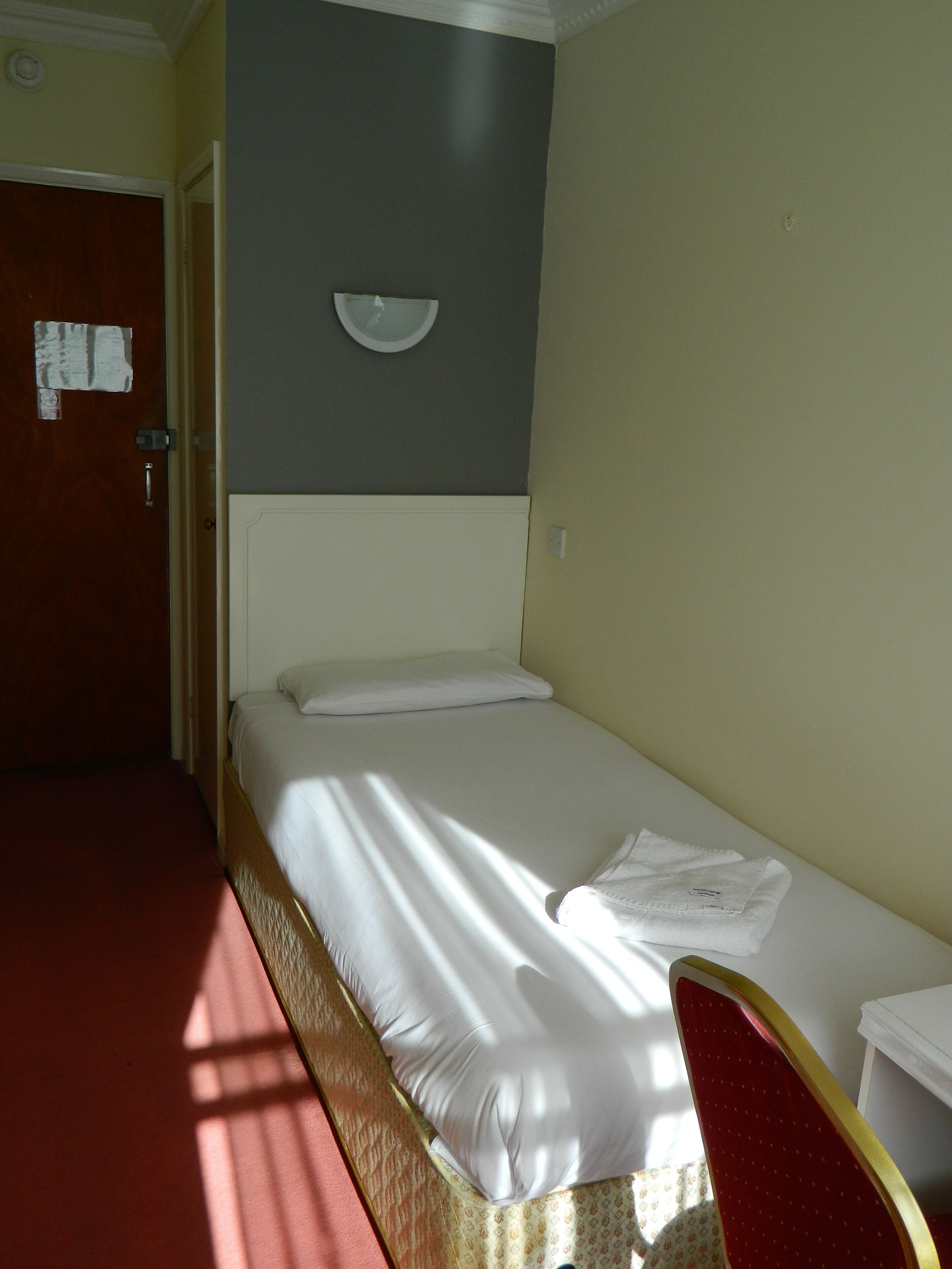 Een enkel bed met witte lakens, een rode stoel en een klein tafeltje met een witte tafelkleed.
