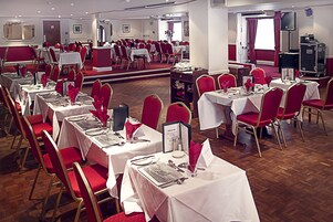 Un salón de banquetes con varias mesas redondas dispuestas para comer, sillas rojas y un escenario para presentaciones.
