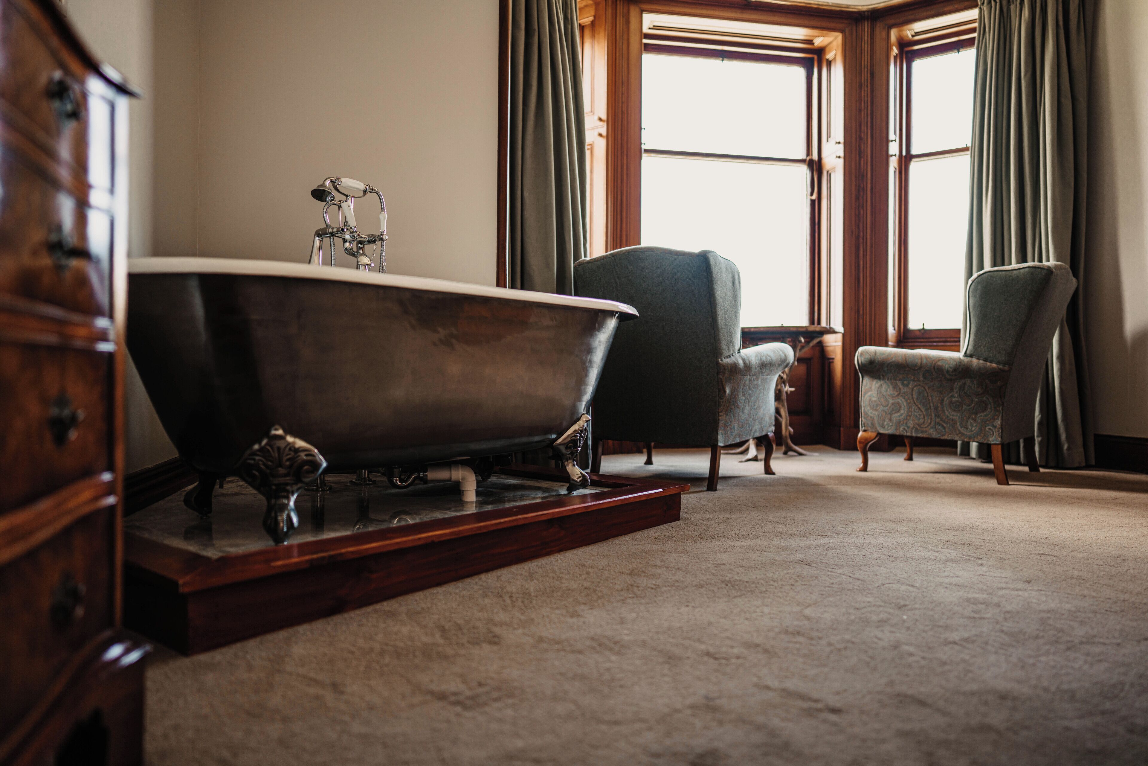 A room with a large bathtub, two chairs, and a wooden dresser.