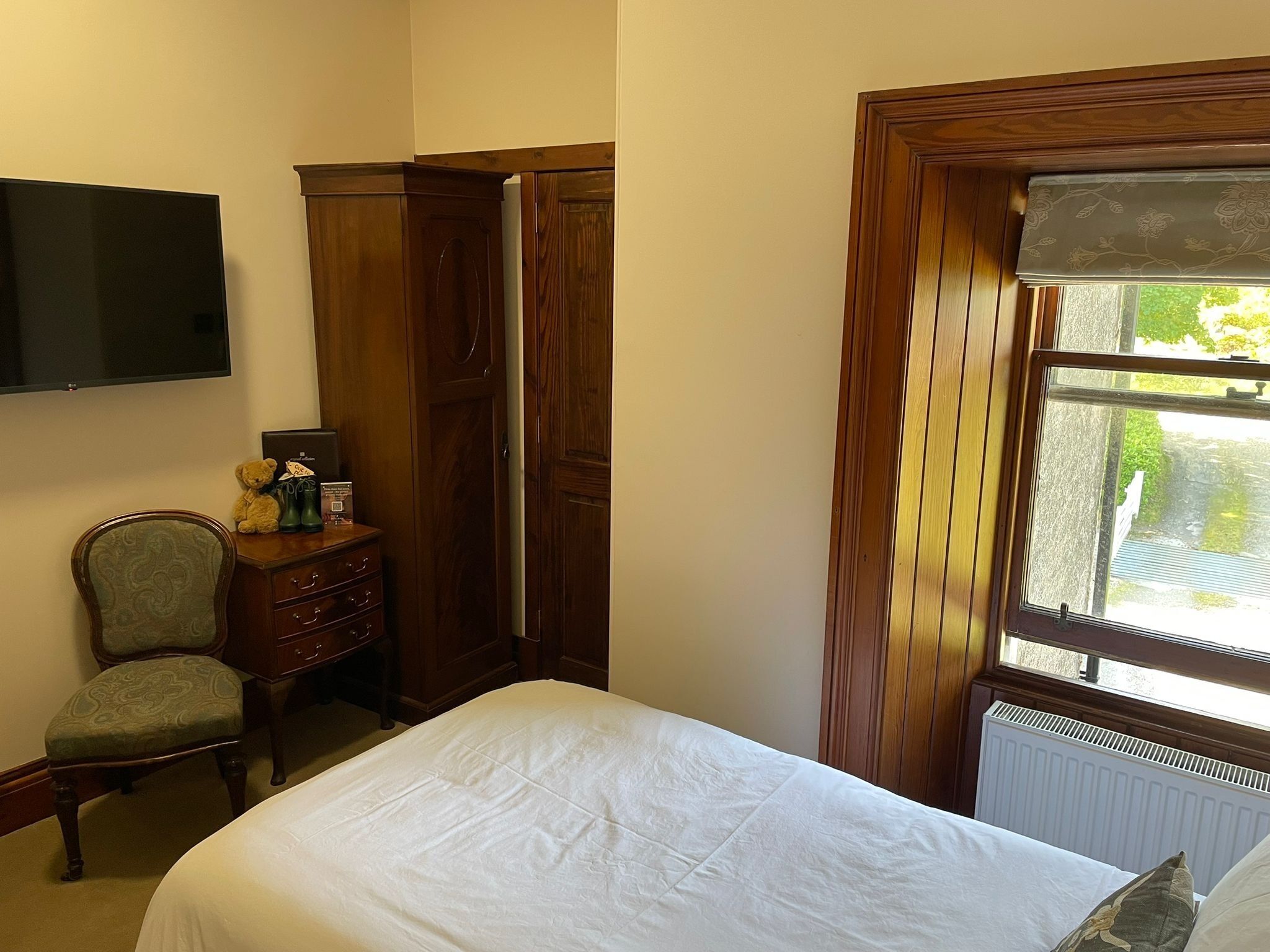 A bedroom with a bed, a dresser, a chair, a television, and a window with a view of greenery.