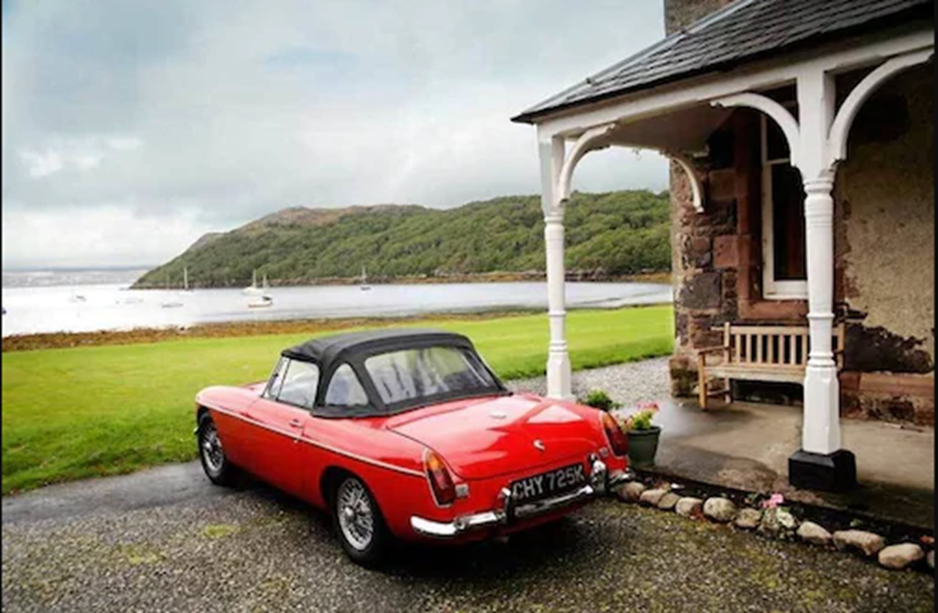 A red convertible parked in front of a stone house with a porch, overlooking a body of water and green hills.